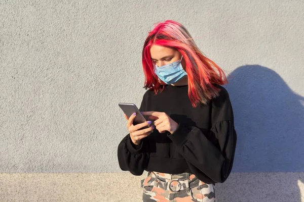 Hipster chica adolescente con máscara de protección médica en la cara, con teléfono inteligente en las manos —  Fotos de Stock