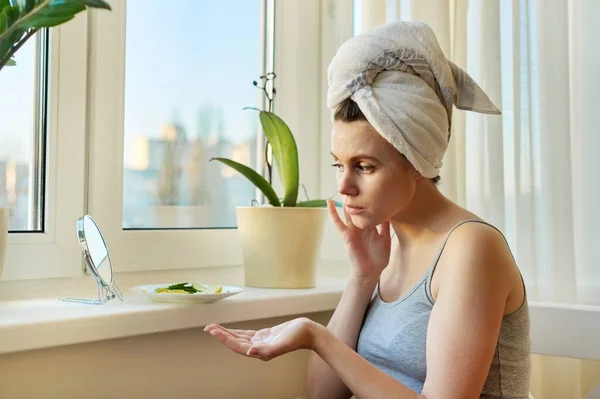 Jovem mulher perto de casa janela olhando no espelho, aplicando creme facial — Fotografia de Stock