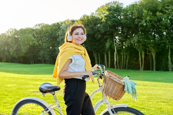 Beautiful happy middle-aged woman in headphones with bicycle in park