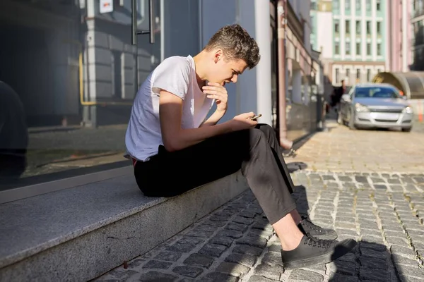 Fiú tizenéves 16,17 éves, okostelefonnal, ül az utcán a nyári város — Stock Fotó