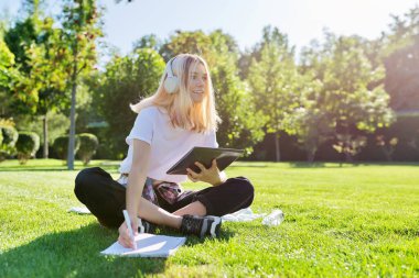 Kulaklıklı, dijital tabletli genç bir öğrenci çimenlikte oturmuş deftere yazı yazıyor.