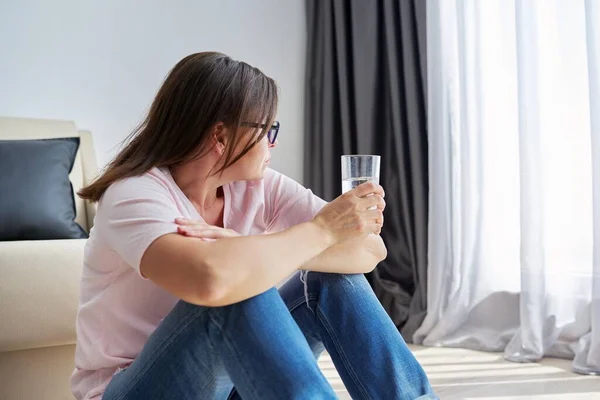 Serious triste de mediana edad madura mujer sentada en casa sosteniendo un vaso de agua — Foto de Stock