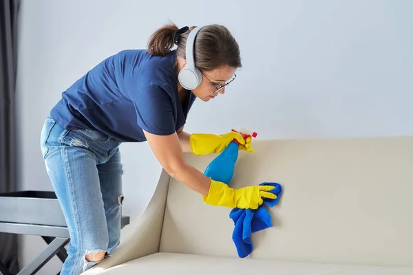 Limpieza del hogar, mujer en auriculares con trapo y spray con detergente lavado sofá de cuero —  Fotos de Stock