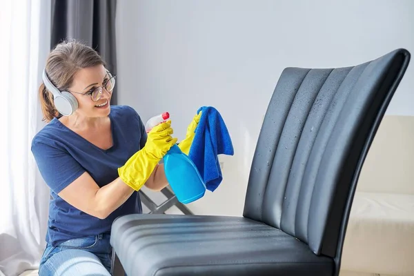 Hemstädning, kvinna i hörlurar och handskar med trasa och spray med tvättmedel läderstol — Stockfoto