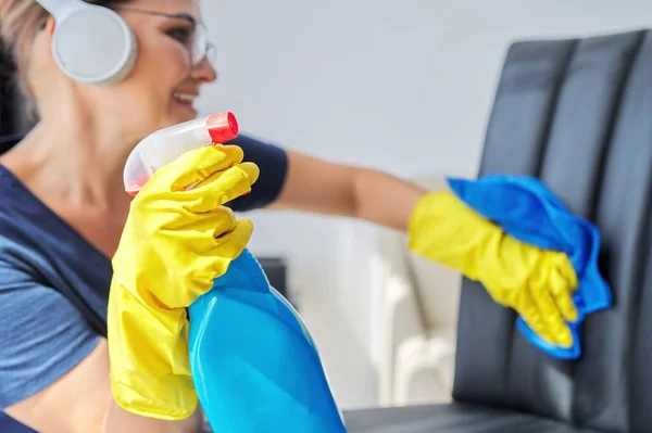 Home cleaning, woman in headphones and gloves with rag and spray with detergent washing leather chair — Stock Photo, Image