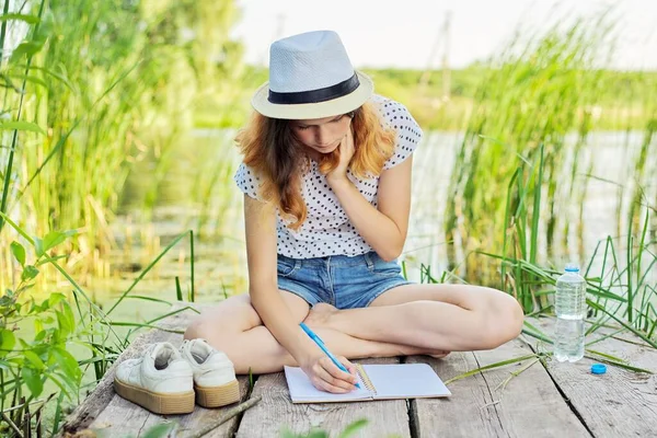 Tvůrčí teenager píše do sešitu, dívka sedí na dřevěném molu u jezera — Stock fotografie