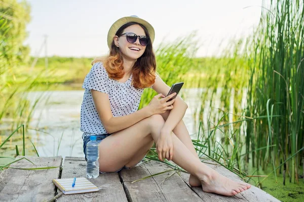 Šťastná dospívající dívka v klobouku s chytrým telefonem na dřevěném molu jezera s rákosím — Stock fotografie