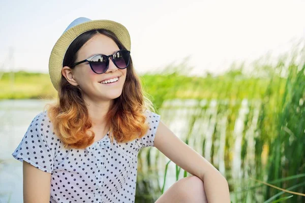 Happy smiling teenager girl in hat sunglasses sitting on the shore of the lake — Stock Photo, Image