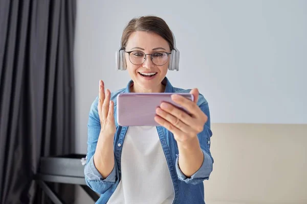 Femme parlant positif mature dans les écouteurs regardant l'écran du smartphone, appel vidéo — Photo