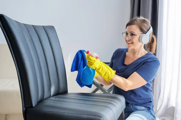 Hemstädning, kvinna i hörlurar och handskar med trasa tvätt läderstol — Stockfoto