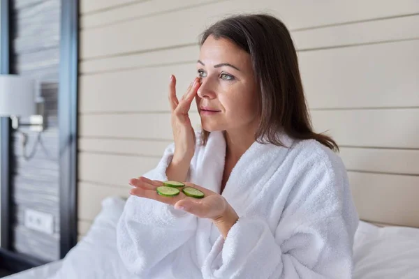 Volwassen vrouw in badjas doen thuis spa behandelingen met komkommers — Stockfoto