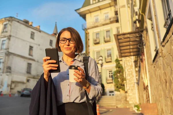 Счастливая зрелая деловая женщина в очках с чашкой кофе со смартфоном на городской улице — стоковое фото