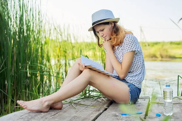 Tiener meisje in hoed leest dagboek zitten op oever van het meer, natuur landschap achtergrond — Stockfoto