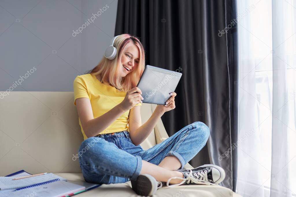 Teenager girl student in headphones looking in digital tablet, sitting at home on couch