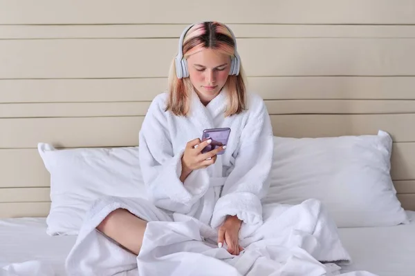 Chica adolescente en albornoz blanco y auriculares con teléfono inteligente en sus manos —  Fotos de Stock