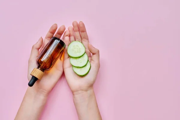 Womans hands with glass bottle with liquid, cucumber slices on pink background — Stock Photo, Image