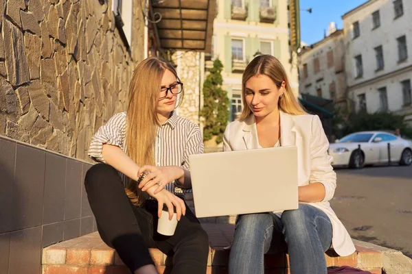 Две молодые улыбающиеся женщины смотрят на монитор ноутбука, сидят на улице — стоковое фото