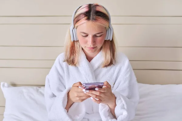 Teenager dívka v bílém županu a sluchátka s chytrým telefonem v ruce — Stock fotografie
