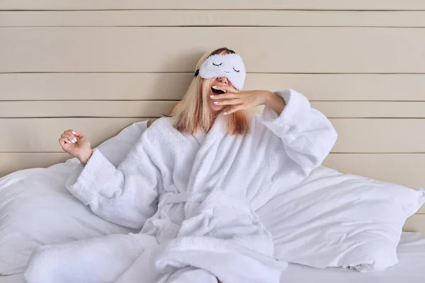 Jovem mulher na cama branca em roupão de banho, máscara ocular, bocejos femininos — Fotografia de Stock