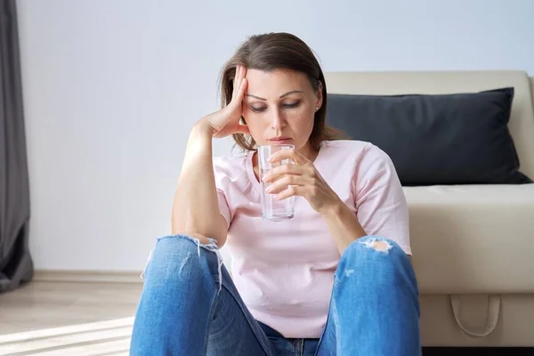 Triste mujer de mediana edad sentada en casa en el suelo con un vaso de agua —  Fotos de Stock
