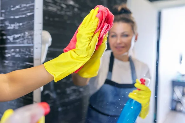 Housewife in apron gloves with detergent washing the mirror in the bathroom — Stock Photo, Image