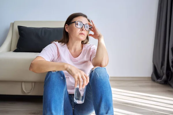 Triste mujer madura infeliz sentada en casa en el suelo con un vaso de agua —  Fotos de Stock