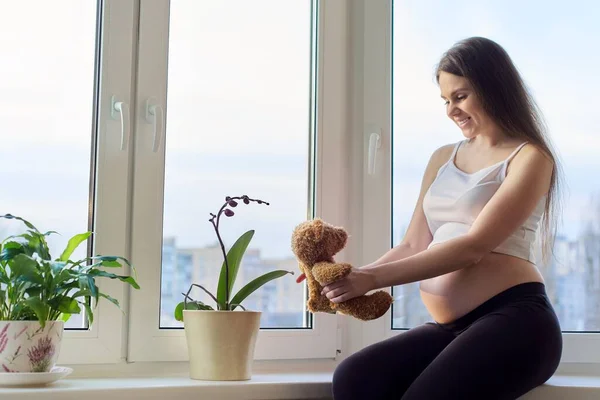 Беременная молодая счастливая женщина с игрушечным плюшевым мишкой, у окна дома — стоковое фото