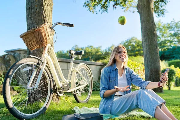 Gelukkig vrouw zitten op het gras gooien appel maken video call op smartphone — Stockfoto