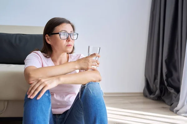 Traurige unglückliche reife Frau sitzt zu Hause mit einem Glas Wasser auf dem Fußboden — Stockfoto