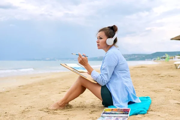 해변의 모래사장에 앉아 바다를 스케치하는 장성 한 여자 — 스톡 사진