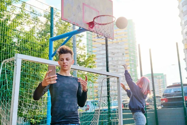 Guy teenager recording video stream using smartphone, on sports street court, girl playing basketball — Stock Photo, Image