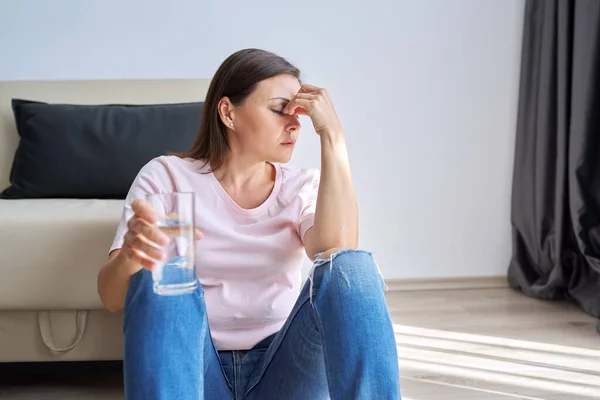 Triste mujer madura infeliz sentada en casa en el suelo con un vaso de agua — Foto de Stock