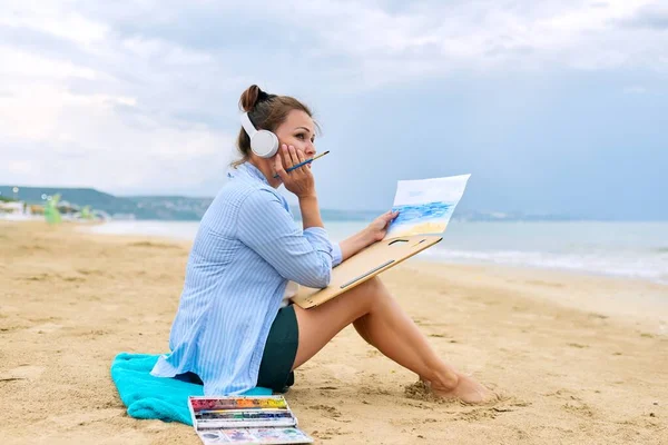 Relajación en el mar, mujer madura en auriculares con acuarelas pintura — Foto de Stock