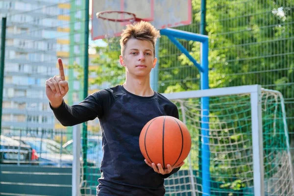 Niño serio adolescente con pelota de baloncesto mostrando el pulgar hacia arriba señal de atención gesto de la mano — Foto de Stock