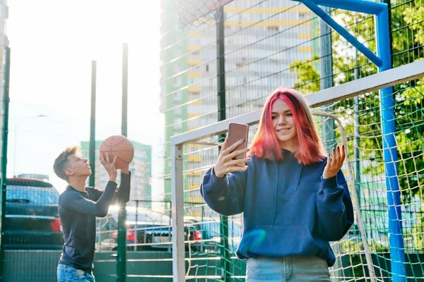 Flicka tonåring inspelning video ström med hjälp av smartphone, på sport street court, kille som spelar basket — Stockfoto