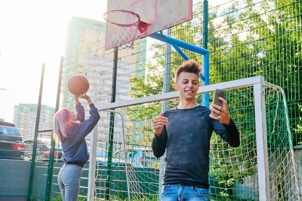 Guy teenager recording video stream using smartphone, on sports street court, girl playing basketball — Stock Photo, Image