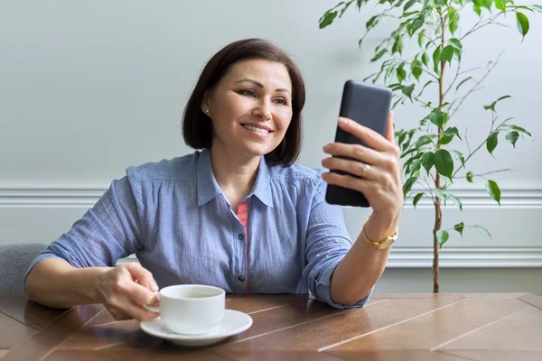 Зріла жінка зі смартфоном сидить вдома посміхаючись дивиться на екран телефону — стокове фото