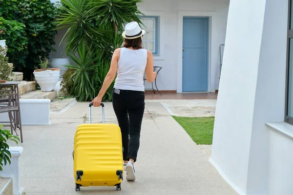Woman with suitcase walking outdoor through territory of resort spa hotel, back view