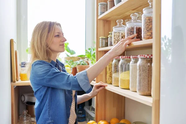 Home kitchen, pantry, woman near wooden rack with food in jars and containers — Stock Photo, Image