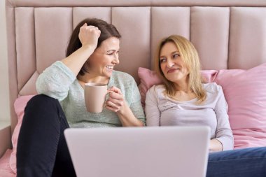 İki yetişkin kadın dizüstü bilgisayara bakıyor ve evde yatakta oturuyor.