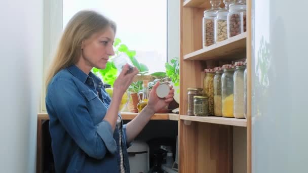 Almacenamiento de alimentos en la despensa, mujer de mediana edad en la cocina cerca de estante de madera con frascos de cereales — Vídeos de Stock