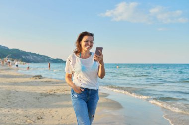 Kumsalda akıllı telefonuyla yürüyen orta yaşlı bir kadın.