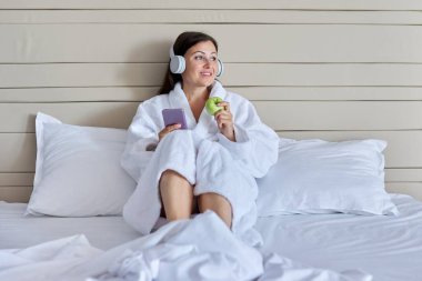Olgun bir kadın, kulaklıkla yatakta, akıllı telefonuyla, yeşil elmayla dinleniyor.