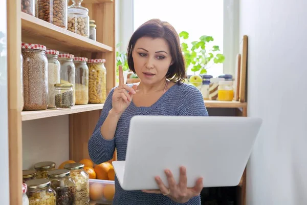 Woman in kitchen pantry with stored products, holding laptop — Stock Photo, Image