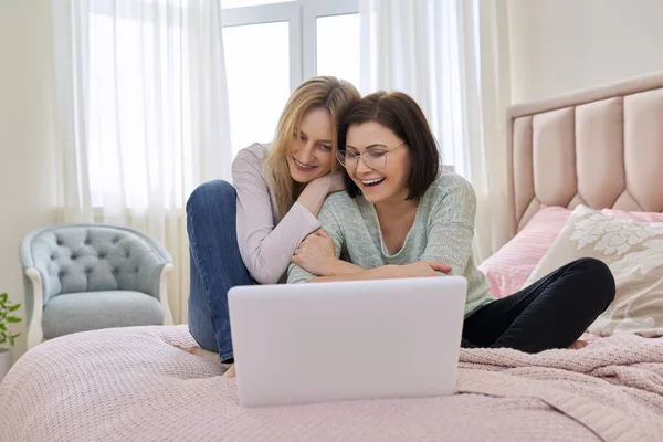 Dos mujeres de mediana edad descansando sentadas juntas en casa en la cama, mirando en la pantalla del ordenador portátil. — Foto de Stock