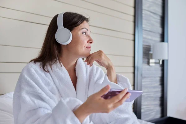 Mujer en albornoz y auriculares escuchando música y relajación, sentada en la cama —  Fotos de Stock