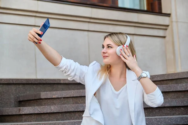 Jovem mulher bonita em fones de ouvido com smartphone descansando sentado em passos na cidade — Fotografia de Stock