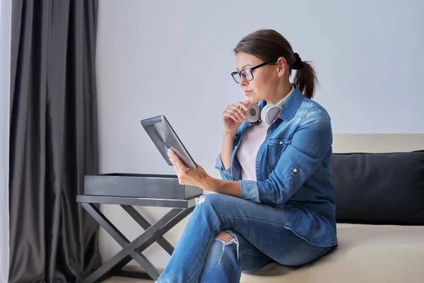 Mujer seria de mediana edad en auriculares con tableta digital en casa en el sofá —  Fotos de Stock