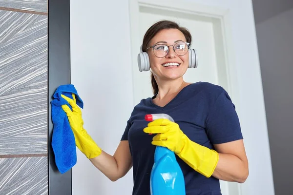 Gammal kvinna i hörlurar handskar med trasa rengöringsmedel hemma — Stockfoto