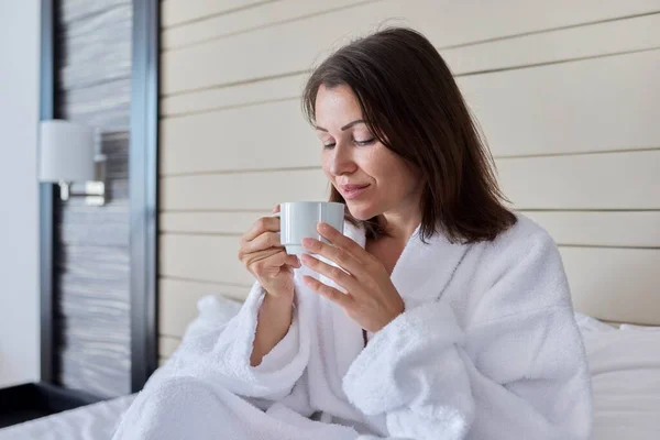 Mulher madura bonita com xícara de café na cama em roupão branco sentado na cama — Fotografia de Stock